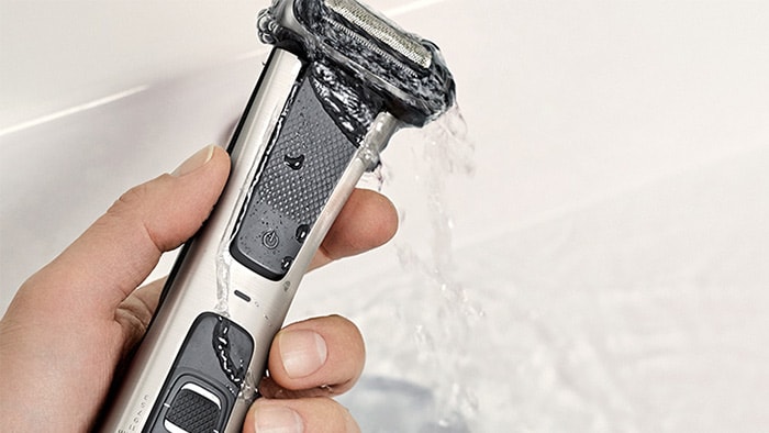 A hand holding a Philips body groomer under a running tap, rinsing it with water.
